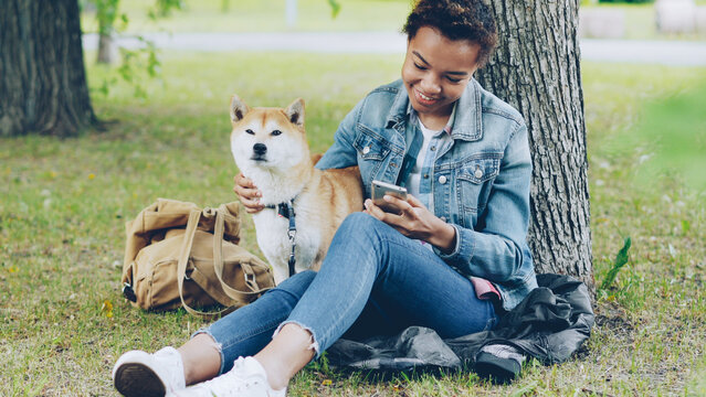 Sociable girl is using smartphone texting friends and stroking her adorable puppy while resting in park at summer weekend. Modern technology, people and animals concept.