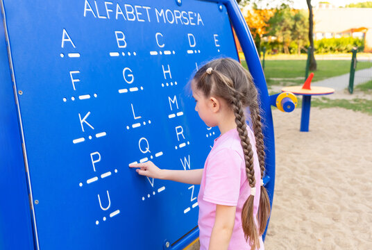 A little girl 7-8 years old with long hair dressed in a pink T-shirt and shorts is studying Morse code on a blackboard in the playground