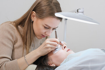 young woman beautician holding tweezers with false lash during lash extension procedure with client. beauty care salon, professional face care. - obrazy, fototapety, plakaty