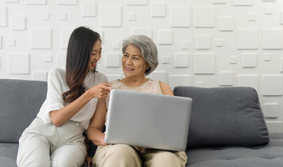 Young asian woman pointing finger at laptop computer monitor, show her grandmother how to surf the Internet. Happy family living together in the living room.