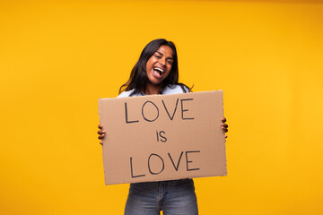 Young indian woman isolated on yellow background holding love is love cardboard poster. Studio shot.