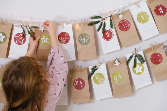 Toddler child gets new advent calendar task, waiting for Christmas. Festive mood for cute curly girl. Kid takes advent bag with dates from the wall at home.