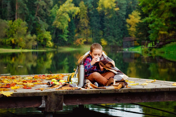 the boy is sitting wrapped in a blanket on a wooden pier next to the lake and drinking hot tea from a thermos. the concept of a picnic in autumn, going out into nature.