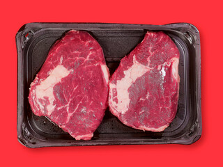 Top quality rib eye steak on a black plastic tray and red color surface background. Premium meat cut product. Butcher craft. Food supply industry.