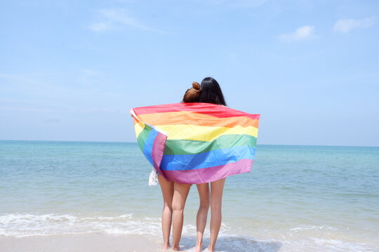 A Lesbian Couple Holding A Rainbow Flag LGBT Symbol On The Beach Against A Bright Blue Sky Background. LGBT Concepts.