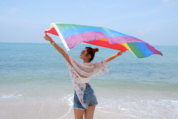 Young woman holding a rainbow flag LGBT symbol on the beach against a bright blue sky background. LGBT Concepts.