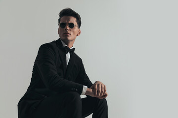 seated cool businessman in tuxedo wearing sunglasses and looking away