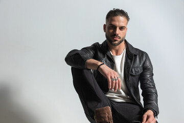 stylish young man wearing black leather jacket posing with elbow on knee