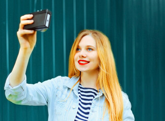 Portrait of happy smiling blonde teenager girl taking selfie with camera in the city