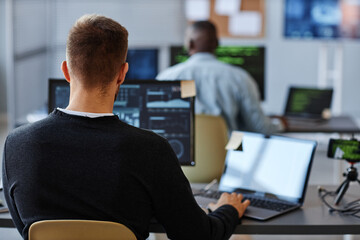 Back view of male programmer or data specialist using computer in office with charts on screen,...