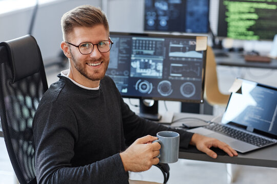 Portrait of young software engineer smiling at camera while posing at workplace in office with coffee cup, copy space