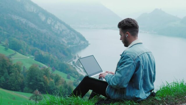 Relaxed male freelancer or student study or work remotely, sitting on the cliff of the mountain in Switzerland. Swiss man using laptop or computer outdoors sitting on green grass, in a foggy day