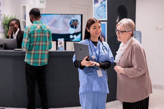 Asian geriatric specialist presenting results of routine tests to elderly caucasian patient. Senior woman looking medical images on computer at doctor office. Hospital admission area with diverse