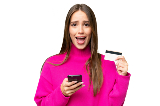 Young beautiful caucasian woman over isolated background buying with the mobile and holding a credit card with surprised expression