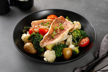 Fillet of fish perch with vegetables, broccoli, cauliflower, tomatoes, potatoes on a dark plate and grey background. Seafood diet plate.