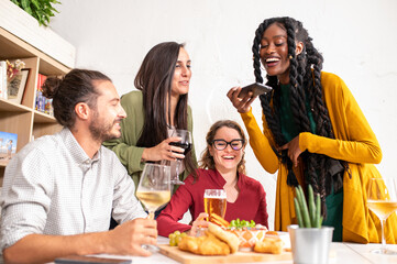 Young African woman smiling while sitting with her friends in a bistro, people taking photos of their meal with her smartphone, influencer enjoying food and drink