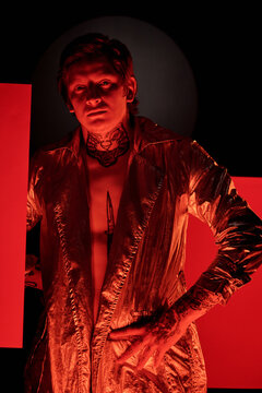 Portrait of young fashion model wearing golden coat on his naked body in tattoos standing in dark neon studio