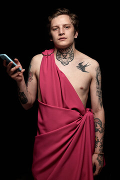 Portrait of young man with tattoo in pink mantle using mobile phone and looking at camera standing against black background