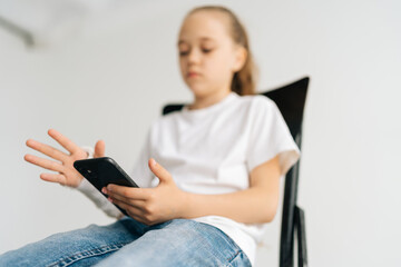 Close-up low-angle view of blonde little girl with broken arm wrapped in white plaster bandage using typing smartphone sitting at chair in light room. Concept of child insurance and healthcare.