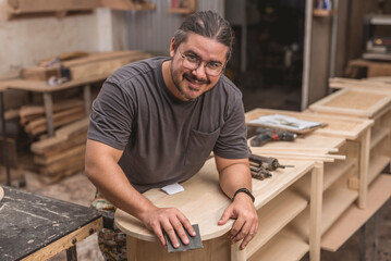 A man smiles at the camera while sanding the edges of a newly built shoe bench at his furniture...