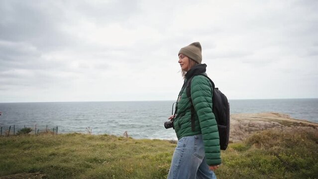 Slow-motion cinematic follow shot of a woman tourist with a backpack and a camera walking along the path with a top view of the sea in the wintertime.