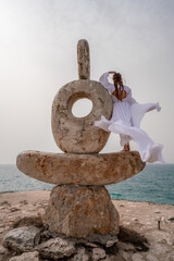 Fototapeta na wymiar A woman stands on a stone sculpture made of large stones. She is dressed in a white long dress, against the backdrop of the sea and sky. The dress develops in the wind.