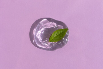 A drop of transparent cosmetic gel or serum with hyaluronic acid with a green leaf on a lilac...