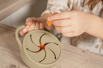 Unrecognizable child taking piece of carrot out from pastel gray silicone snack cup near lid at wooden table. Baby accessories, tableware, first feeding concept