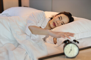 Happy young asian woman in white lingerie lying in bed, wake up late and overslept morning, trying to stop alarm clock. Cute girl look comfortable, need more sleep. Difficult morning get up concept