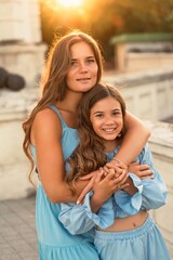 Portrait of mother and daughter in blue dresses with flowing long hair against the backdrop of sunset. The woman hugs and presses the girl to her. They are looking at the camera.