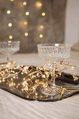 Glass champagne glasses on a handmade tray with lights from a garland, stand on the bed for the new year. Christmas and celebration .