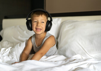 Little sleepy boy sitting in bed wearing wireless headphones and listening to music