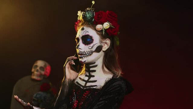 Lady of dead talking on phone call with smartphone, wearing flowers crown and cavalera catrina halloween costume. Woman dressed as santa muerte to celebrate mexican culture holiday. Handheld shot.
