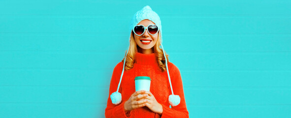 Winter portrait of happy smiling young woman with coffee cup wearing red knitted sweater, white hat with pom pom, heart shaped sunglasses on blue background