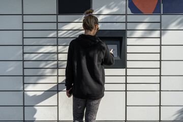 Woman picking up a package from a smart electronic steel parcel locker box, automatic mailboxes. Paczkomat delivery service, collection machine. Using app on her phone to open the door.