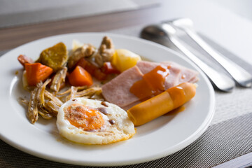 breakfast cuisine with fried egg pepper sausage ham crispy fish steamed pumpkin carrots and baked potato with chili sauce on white dish and placemat with spoon and fork for clean and healthy food