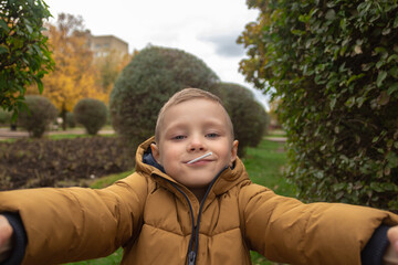 Adorable kid boy in a light brown coat  is taking a selfie outdoor