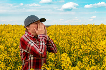 Female farmer with pollen allergy sneezing in blooming rapeseed field. Farm worker wearing plaid...