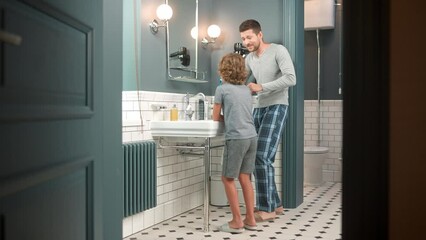 Cheerful Caucasian family father and little teen child brushing teeth in bathroom and dancing singing in good mood. Young happy son with dad having fun while wash together near sink in the morning