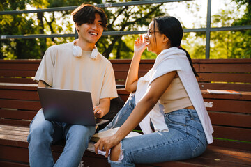 Two multiracial friends laughing while using laptop together in skate park