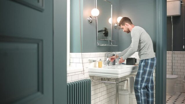Young handsome happy man wearing pajamas standing in bathroom at home and start washing taking toothbrush. Handsome Caucasian guy indoors washing in sink looking at mirror in evening