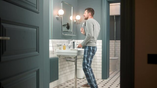 Caucasian happy positive young adult man dancing and singing in bathroom while brushing teeth with toothbrush. Handsome male in pajamas brush his teeth and performing funny dance. Hygiene rituals
