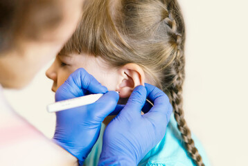 A charming little girl is undergoing the procedure of ear piercing by a professional medical...