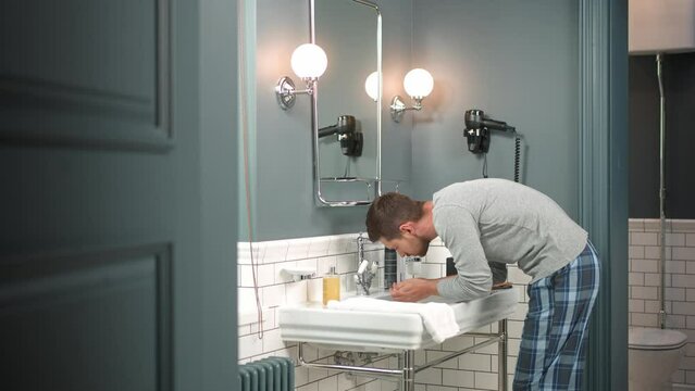 Side view of Caucasian adult man washing his face in bathroom in morning. Handsome male in pajamas cleansing face in sink at home. Healthy cleansing routine. Personal hygiene. Through the door shot