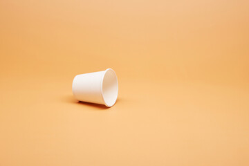 Disposable white single one recyclable cardboard paper cup knocked over the surface isolated on the bright solid fond plain sandy beige background