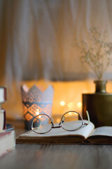 Plate of chocolate chip cookies, stack of vintage books, reading glasses, cup of tea or coffee, lit candle and fairy lights. Hygge at home. Selective focus.