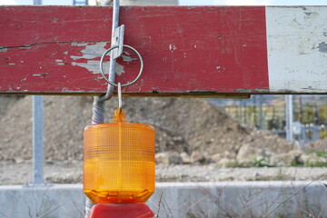 View on the wireless warning construction light of beacon shape and orange color hanging from wooden red and white construction barrier. Behind is building ground with heap of earth and clay.