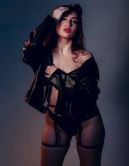 Plus size woman in black leather jacket. Beautiful curvy body, seductive lady posing In underwear. Attractive oversized female In lingerie standing on dark blue studio background.