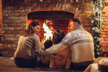 Happy young family parents with two small kids gathering around brick fireplace in cozy and warm country house during Christmas holidays
