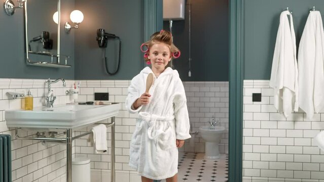 Pretty Cute Caucasian Little Girl In White Robe And With Curlers On Hair After Bath Dancing In Bathroom Holding Hair Brush In Hand. Small Child Dance Indoors Looking At Camera And Smiling. Leisure